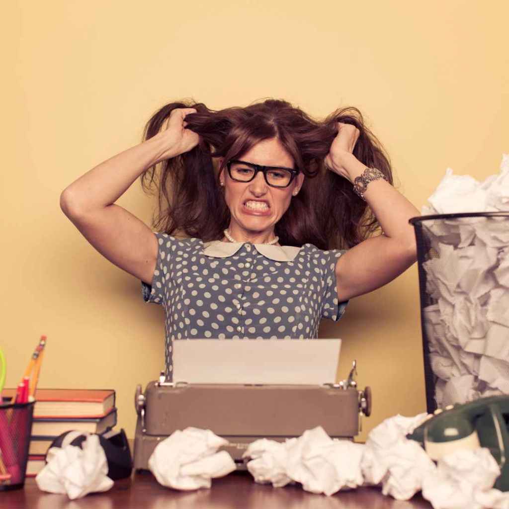 writer pulling her hair and sitting in front of a type writer on a desk with a trash can next to her...frustrated writer